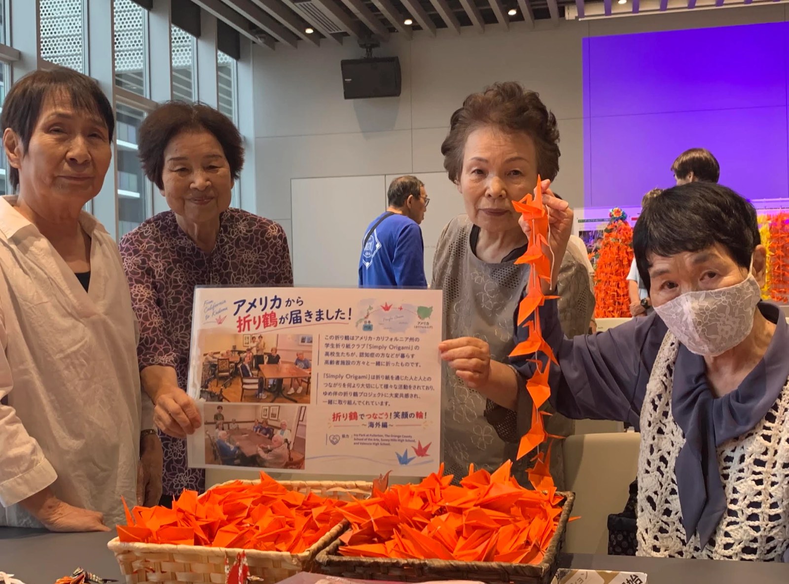 Four women in Kadoma, Japan hold a sign reading “Cranes have arrived from America!” beside baskets of orange paper cranes