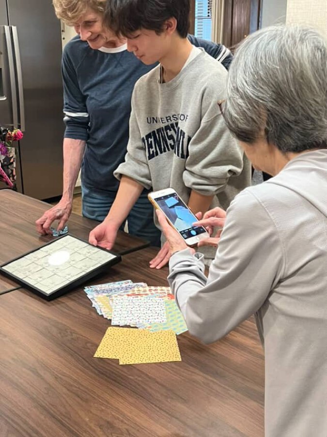 A student volunteer shows origami paper to two senior residents at a kitchen table